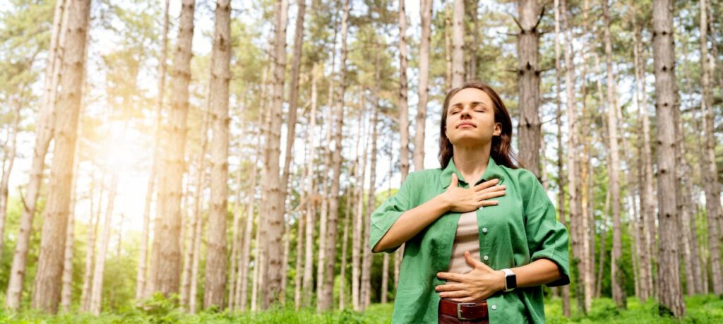 Frau steht im Wald, legt Haende auf Brust und Bauch und praktiziert bewusste Atmung im Stil einer Breathwork Class.